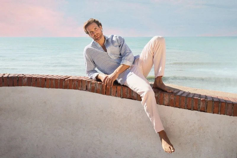 Man in blue printed shirt and pink pants reclining on brick wall in oceanfront setting under pastel sky, lifestyle fashion shot.