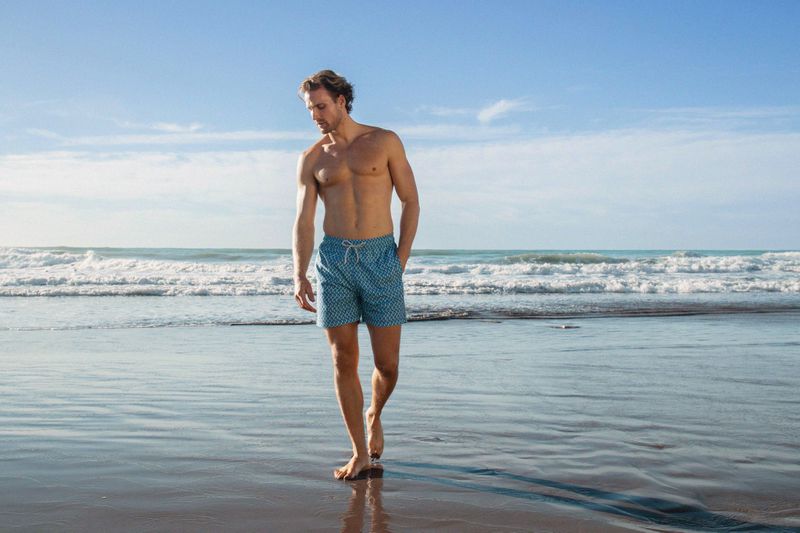 Adult male walking barefoot on a wet sandy beach wearing blue patterned swim trunks, ocean waves and blue sky background on sunny day.