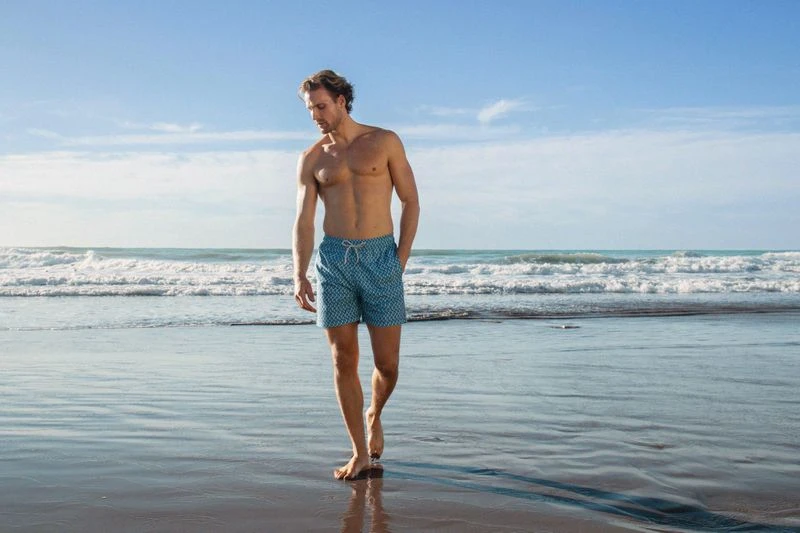 Adult male walking barefoot on a wet sandy beach wearing blue patterned swim trunks, ocean waves and blue sky background on sunny day.