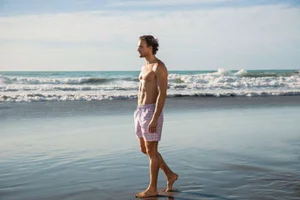 Model walking along the shore of a sandy beach wearing patterned swim shorts against ocean waves