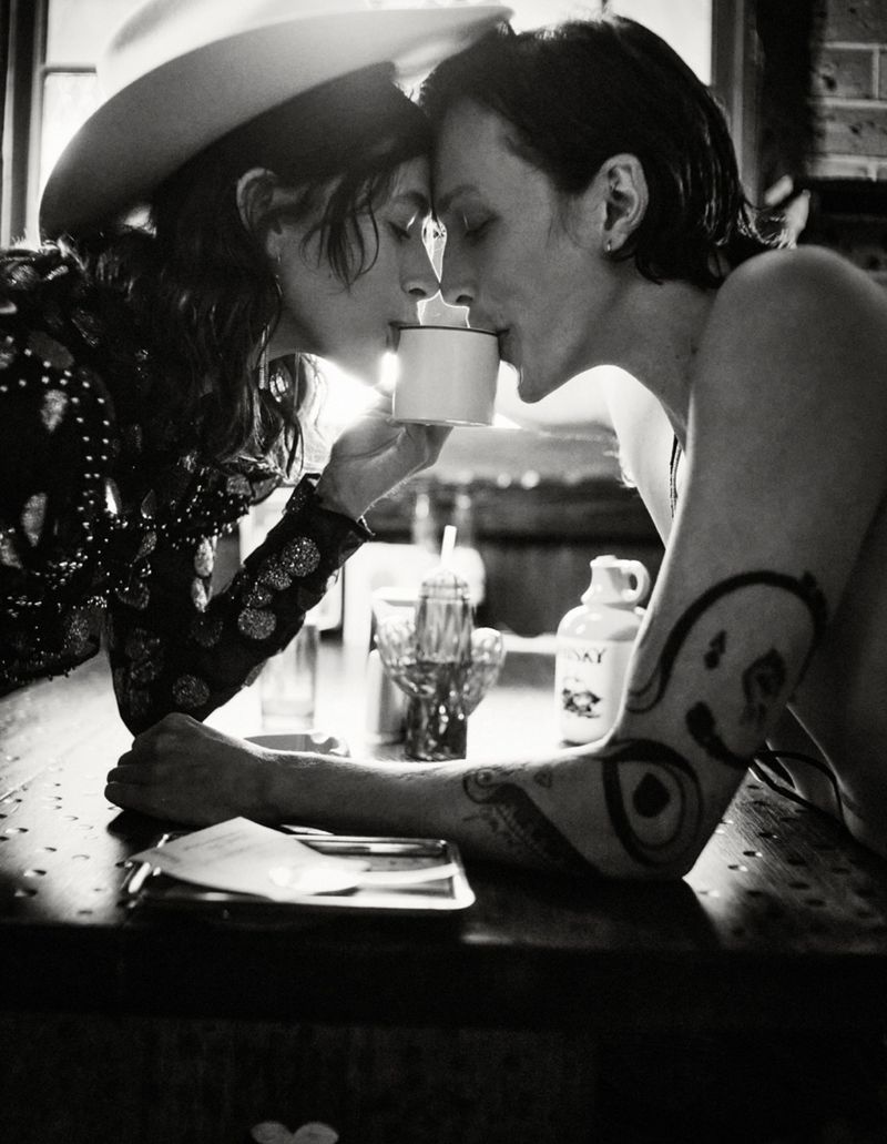 Black and white portrait of two individuals sharing a beverage, one wearing a hat, tattoos visible, in a bar setting with dim lighting.