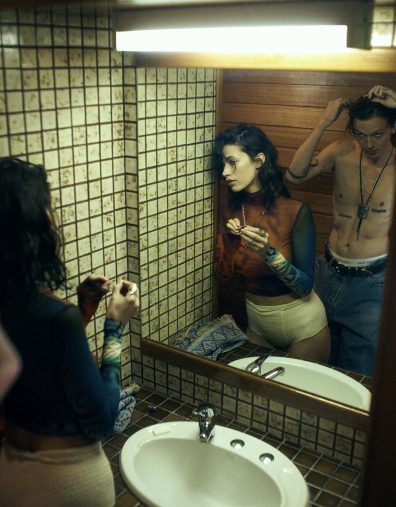 Woman looking in a bathroom mirror with a man standing nearby, both partially visible in a close-up shot. Bathroom features tile and a sink.