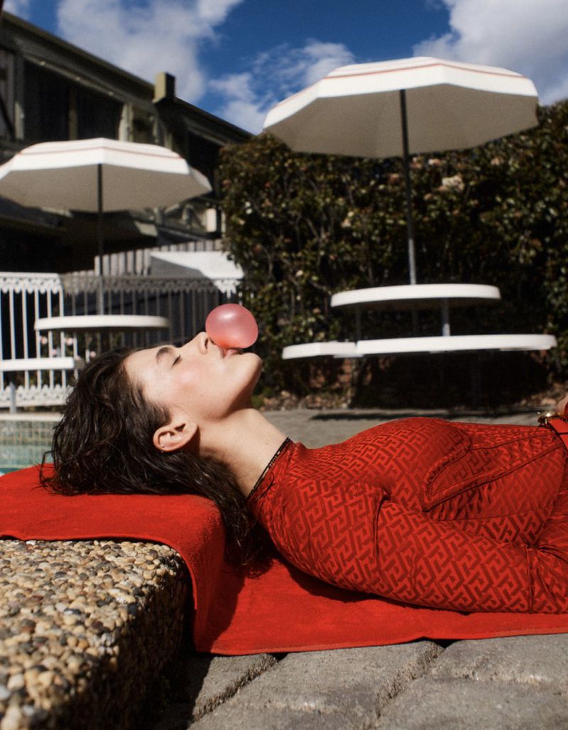 Woman in red patterned dress lying on a red towel, blowing bubble gum bubble, outdoor lounge, sun umbrellas, and building.