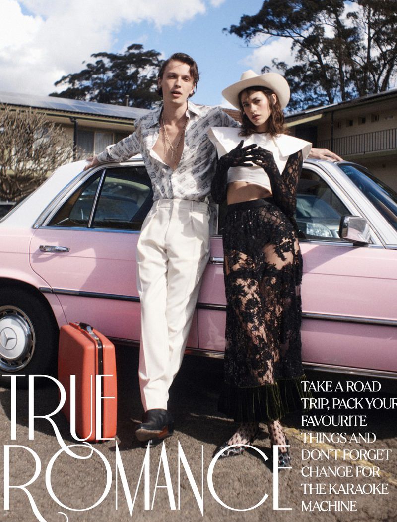 Editorial fashion shot of a man and a woman in front of a pink car with luggage, set against a suburban house and tree-lined sky.