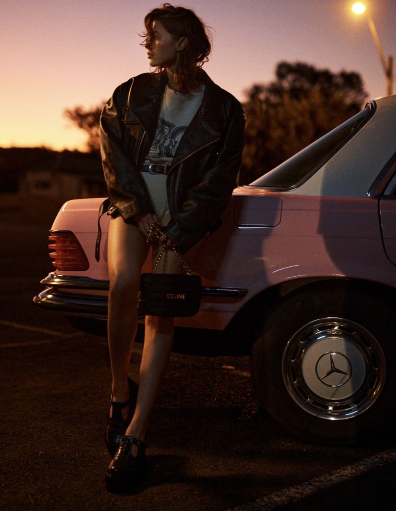 Woman in a leather jacket poses next to a pink car at dusk, with a black purse and dark shoes, on an asphalt parking lot.