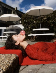 Woman lying poolside on a red towel blowing a pink bubblegum bubble under white patio umbrellas