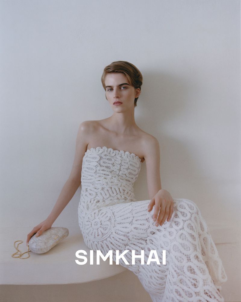 Woman in a white lace gown sits against a wall, holding a decorative clutch, fashion photoshoot, indoor setting.