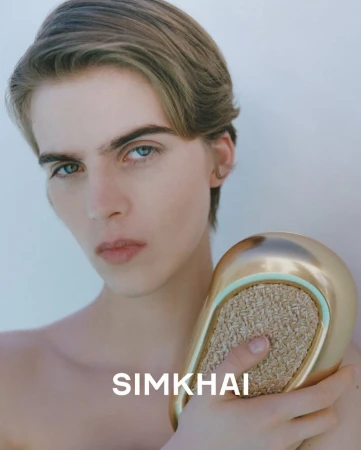 Close-up of a fair-skinned male model with blond hair and blue eyes holding a gold-colored clutch in a white background.