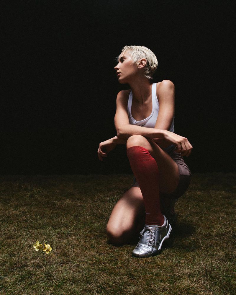 Woman in athletic wear poses on a grass field next to a yellow flower, with a dark background and silver soccer cleats.