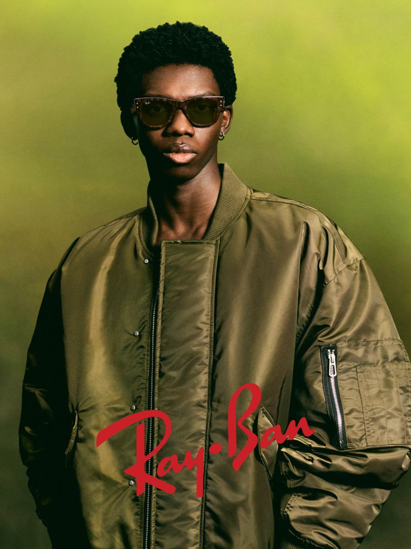 A fashionable adult male model of African descent poses in a stylish olive bomber jacket and sunglasses against a blurred green backdrop.
