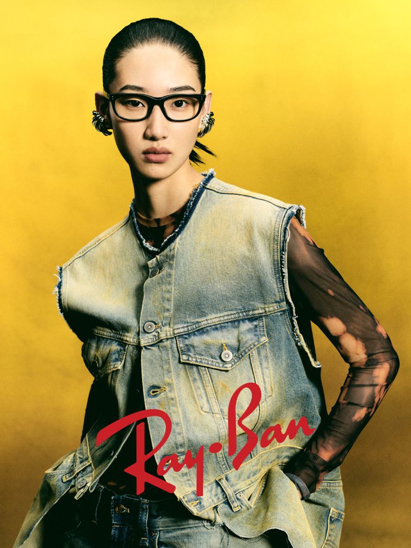 Model wearing black Ray-Ban glasses and denim vest against yellow background