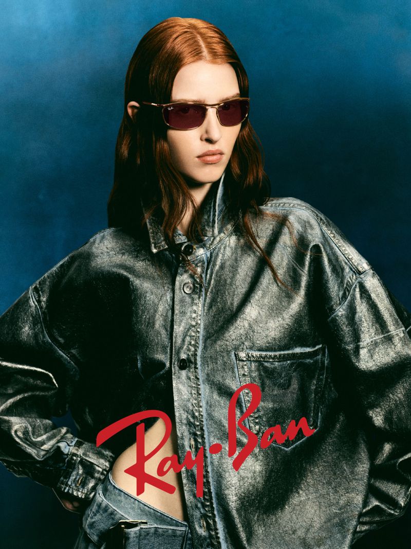 A fair-skinned woman with red hair, wearing sunglasses and a denim outfit, poses against a blue studio backdrop with the Ray-Ban logo.