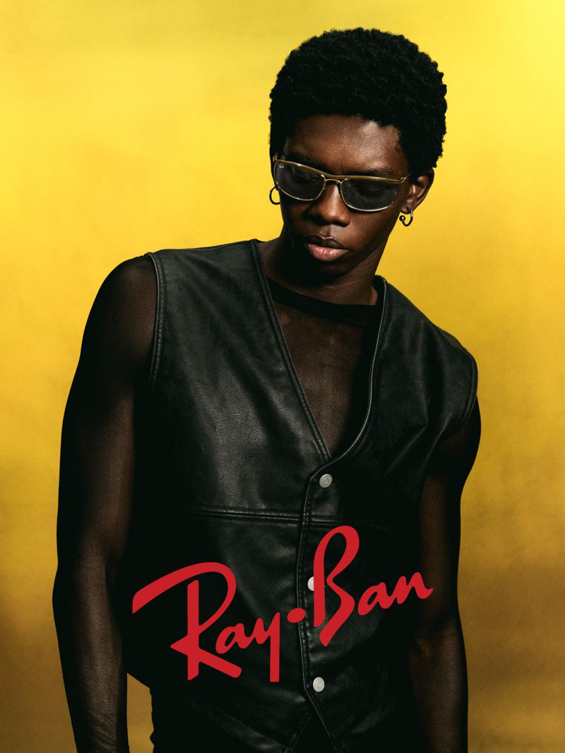 Portrait of a Black man wearing sunglasses and a black vest against a yellow backdrop with the Ray-Ban logo at the bottom of the frame.