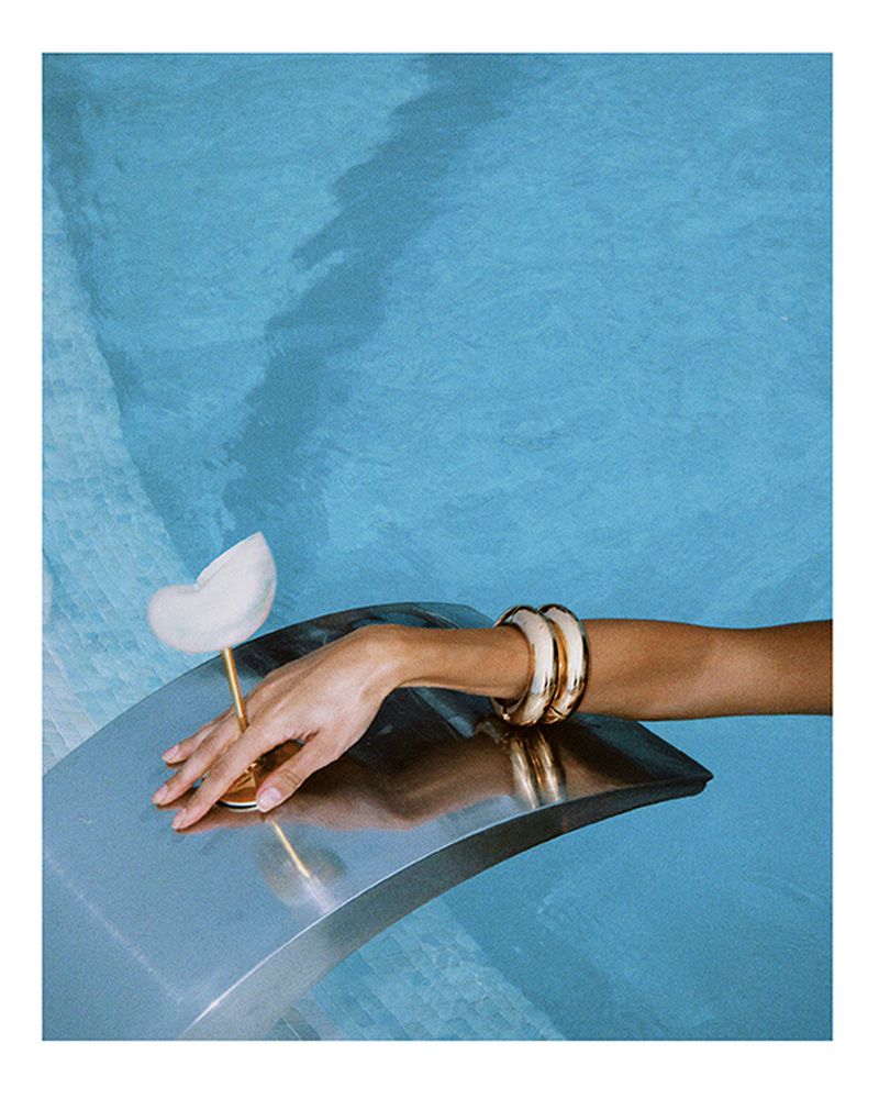 Hand with gold bracelets and a sculptural glass on a silver surface, against a blue patterned background. Fine art photography.