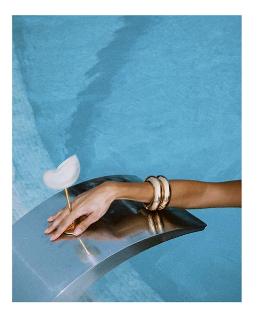 Hand with gold bracelets and a sculptural glass on a silver surface, against a blue patterned background. Fine art photography.