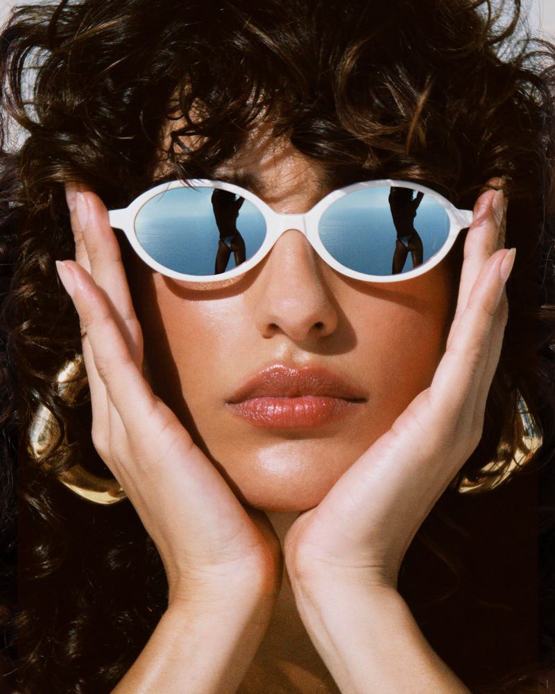 Close-up portrait of a woman wearing white sunglasses with reflective lenses, holding her face with her hands.