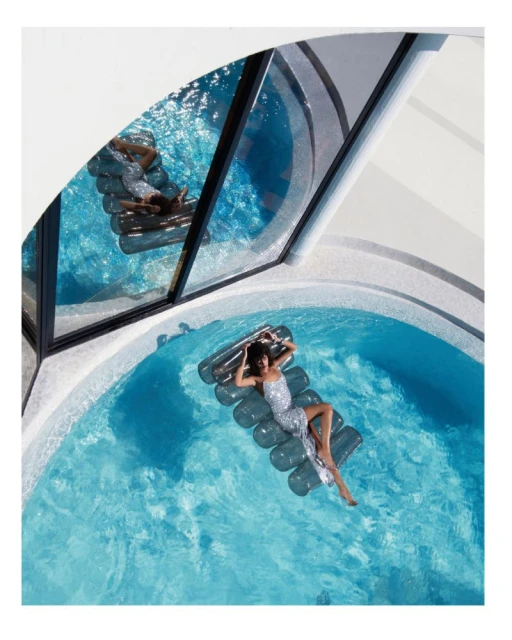 Woman in sequin dress relaxes on inflatable raft in a turquoise pool with bright sunlight reflecting off of the water.