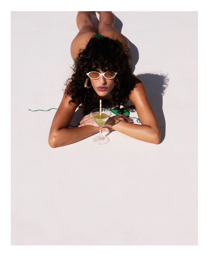 Woman in a bikini lying on a white surface, drinking from a clear martini glass with a straw, wearing sunglasses and earrings.