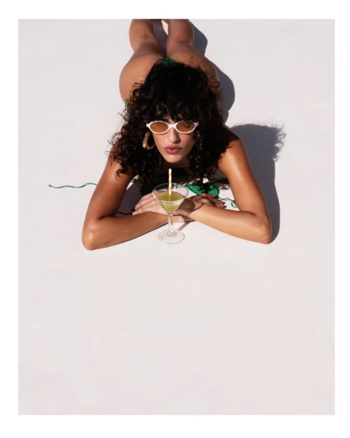 Woman in a bikini lying on a white surface, drinking from a clear martini glass with a straw, wearing sunglasses and earrings.