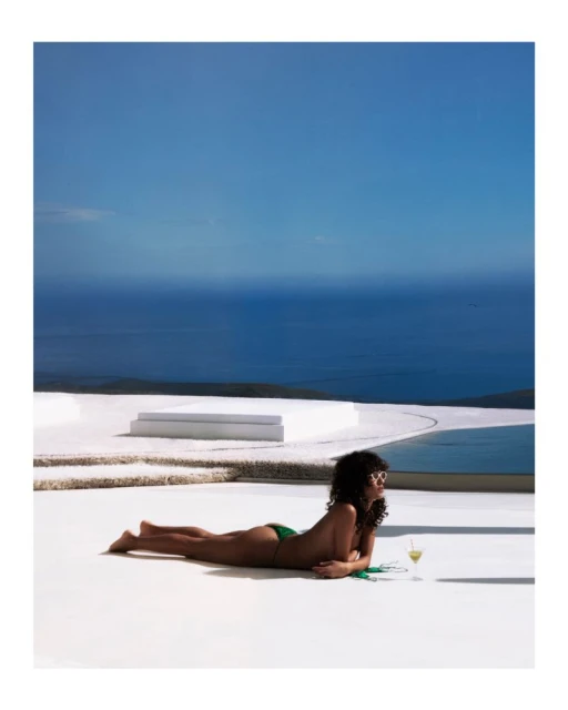 A woman in a bikini lying on a white terrace with a drink, with the ocean and sky visible in the background on a sunny day.