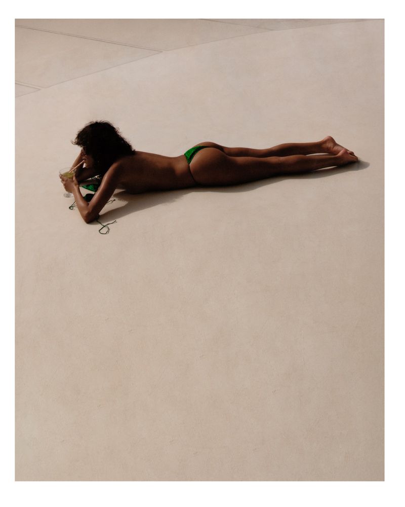 Adult female lying on her stomach on a cream-colored surface, wearing a green bikini bottom and holding a green object near her face.