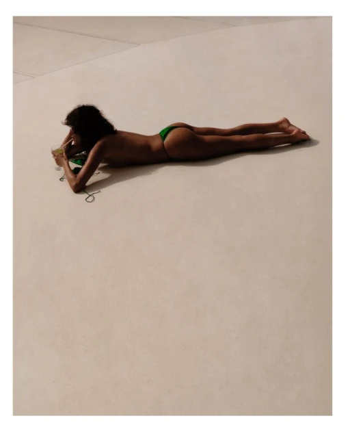 Adult female lying on her stomach on a cream-colored surface, wearing a green bikini bottom and holding a green object near her face.