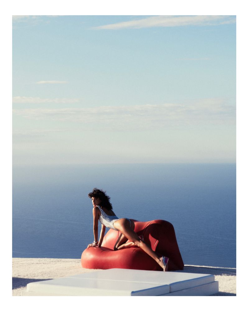 Woman in swimsuit posing on red beanbag chair, outdoor setting with ocean and sky backdrop, creating a summery and stylish image.