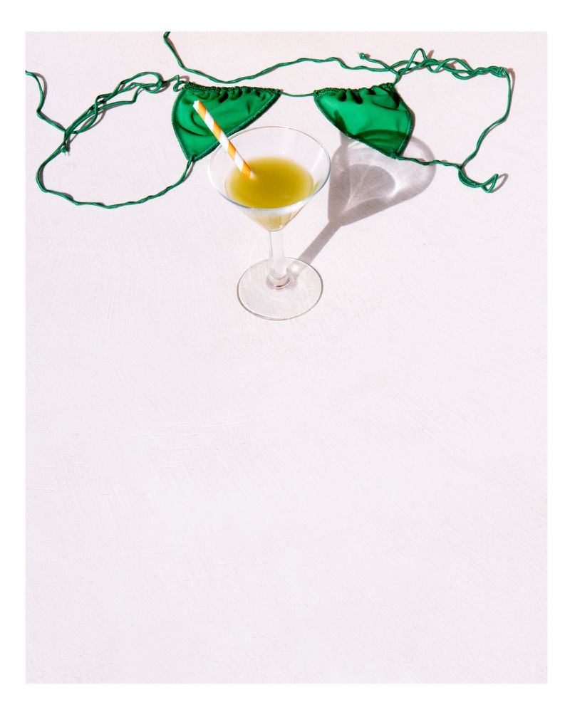 Bright studio shot featuring a clear martini glass filled with a yellow liquid next to a green swimsuit top on a white surface.