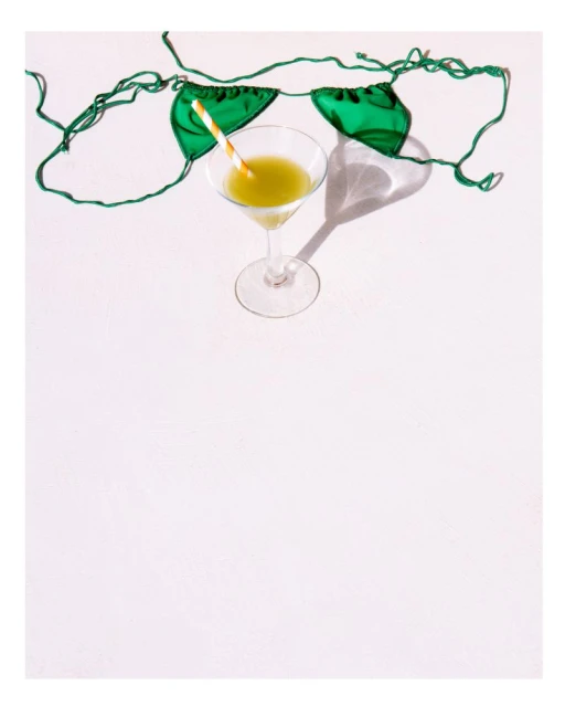 Bright studio shot featuring a clear martini glass filled with a yellow liquid next to a green swimsuit top on a white surface.