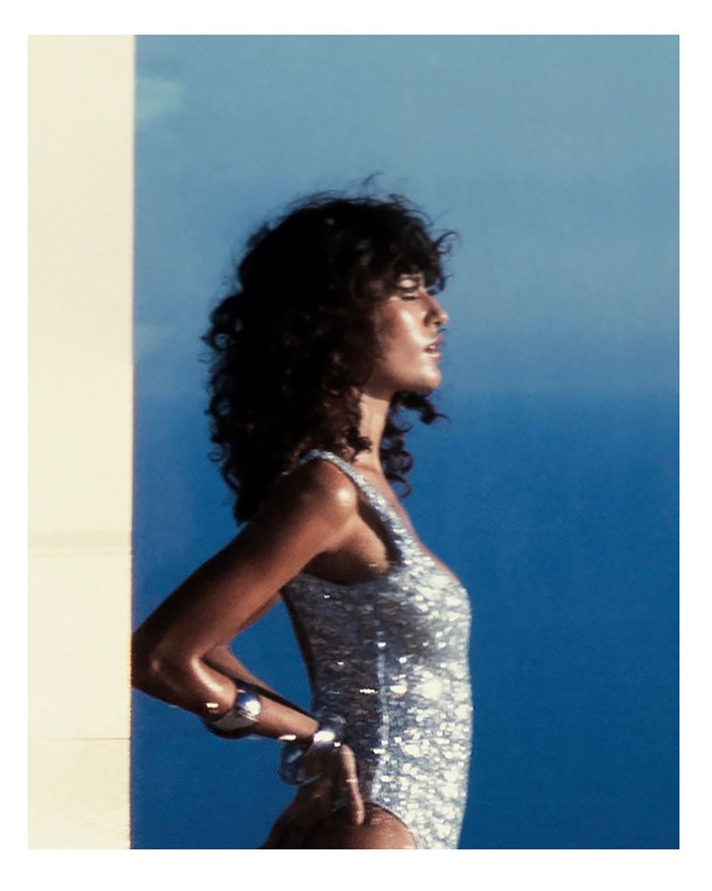 A woman in a shimmering swimsuit poses in front of a blue and white background. The photo features a profile shot, highlighting her jewelry.
