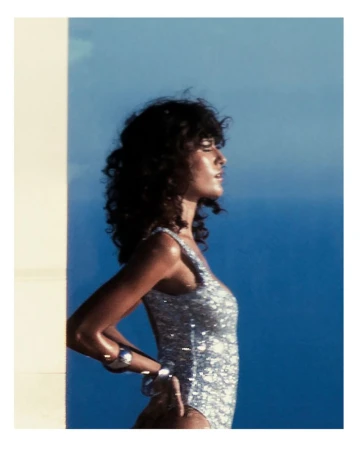 A woman in a shimmering swimsuit poses in front of a blue and white background. The photo features a profile shot, highlighting her jewelry.