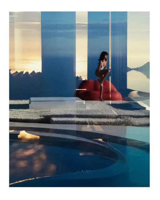 Woman in white swimsuit sits on a large red lip-shaped seat with an ocean and coastal background visible through glass panels.