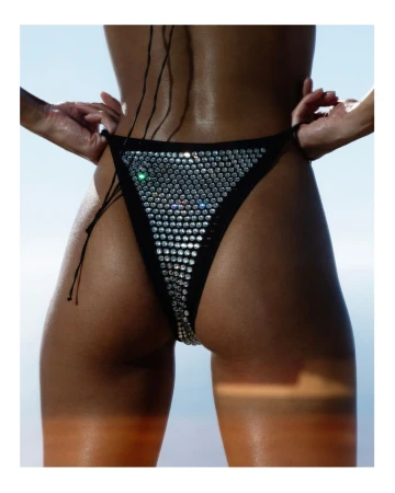 Close-up back view of a tanned adult female with a black bikini bottom covered in rhinestones, set against a bright sky background.