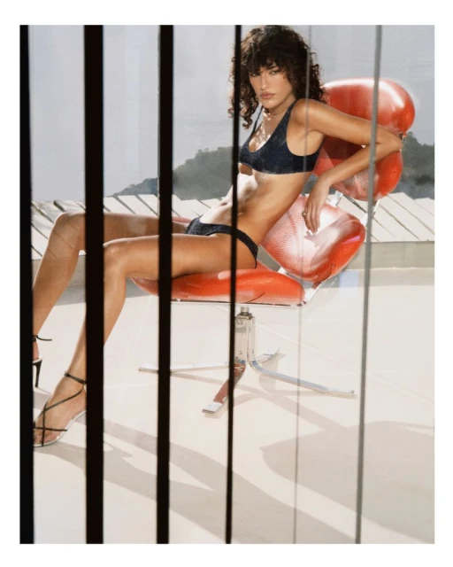 Brunette woman in swimwear poses in a retro red chair against a backdrop of buildings and sky, with vertical lines in the foreground.