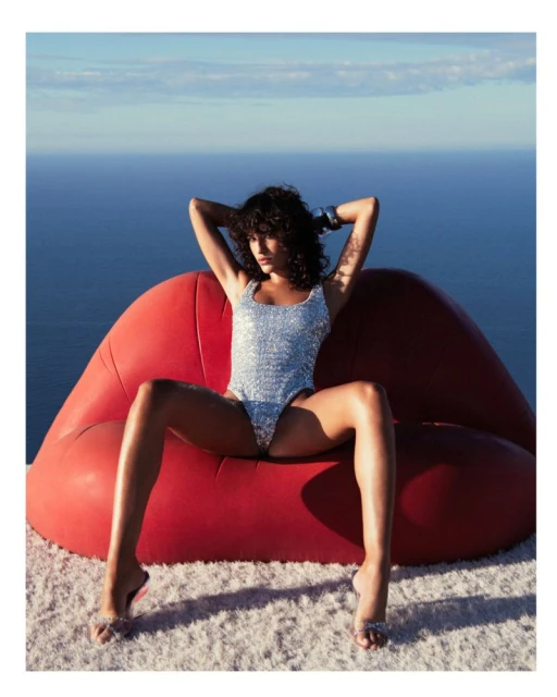 Woman in a sequin bathing suit sits on a red lips-shaped sofa on a white rug against a blue sea and sky background on a sunny day.