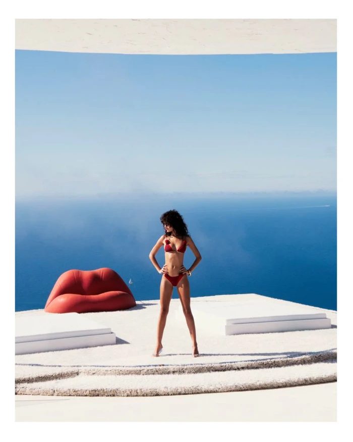 Model wearing a red bikini standing on a white terrace overlooking the blue sea with a red lip-shaped sofa