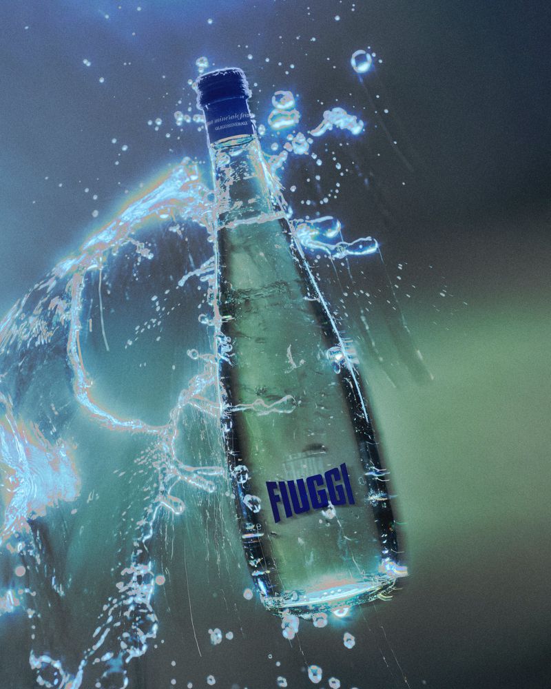 Glass bottle of Fiuggi mineral water with a blue cap, surrounded by water splashes, against a blended blue and green background.