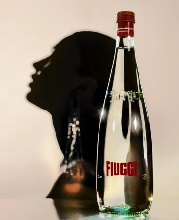 A bottle of Fiuggi mineral water stands in front of a shadowy silhouette of a woman's profile, highlighting the product's refined presentation.
