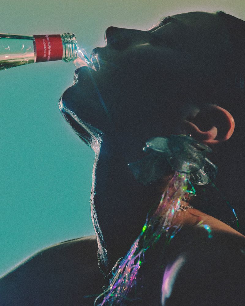 Person drinking water from a bottle. Close-up view of water pouring over face, shoulder and neck. The background is teal color.