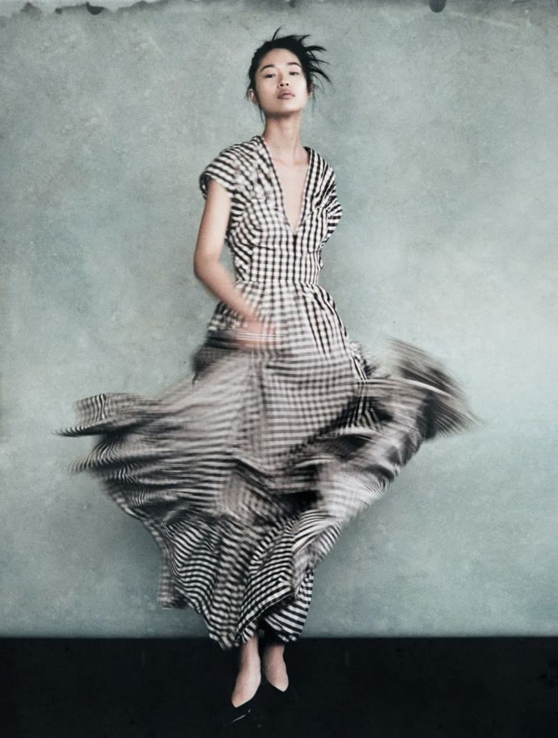 Fashion shot of a woman in a checkered dress with a blurred skirt. She is posing in front of a textured grey wall in a studio setting.