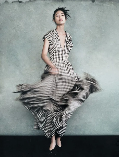 Fashion shot of a woman in a checkered dress with a blurred skirt. She is posing in front of a textured grey wall in a studio setting.