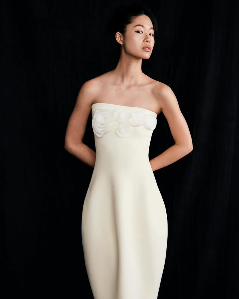 Model wearing a strapless white dress with floral appliques against a dark background for Lela Rose