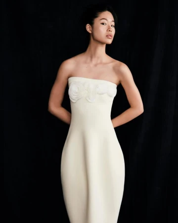 Model wearing a strapless white dress with floral appliques against a dark background for Lela Rose