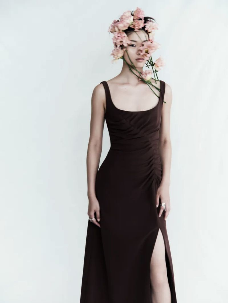 A stylish young woman with an elegant brown dress and a floral headpiece poses against a clean, white backdrop in a fashion-forward portrait.