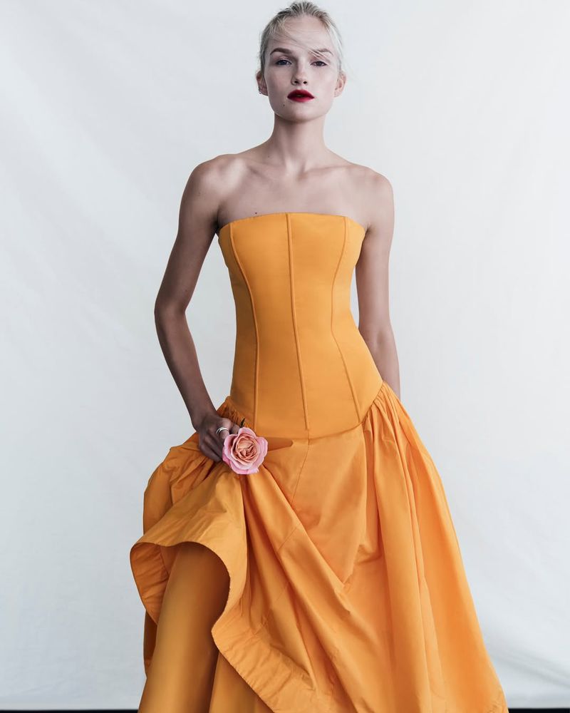 A fair-skinned woman in a strapless, yellow gown holds a pink rose, posing in a full shot against a bright white backdrop in a fashion editorial.
