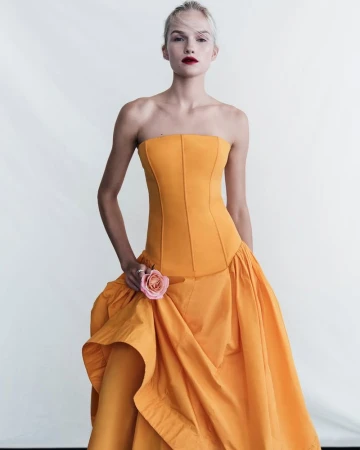 A fair-skinned woman in a strapless, yellow gown holds a pink rose, posing in a full shot against a bright white backdrop in a fashion editorial.