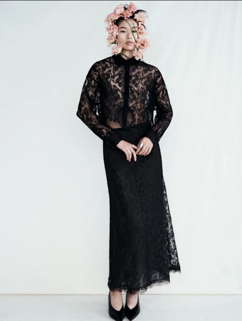 Elegant Asian woman in a sheer black lace top and skirt stands in a studio with a floral headpiece, posing for a fashion shot.