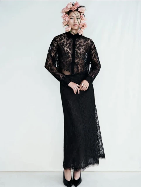 Elegant Asian woman in a sheer black lace top and skirt stands in a studio with a floral headpiece, posing for a fashion shot.