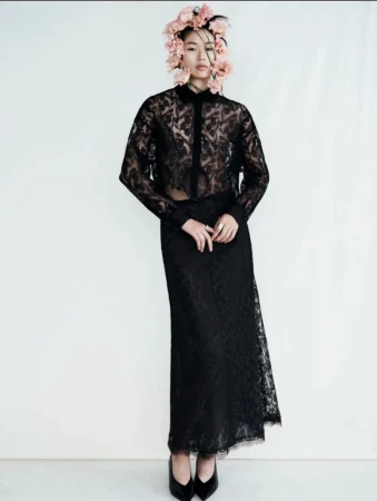 Elegant Asian woman in a sheer black lace top and skirt stands in a studio with a floral headpiece, posing for a fashion shot.