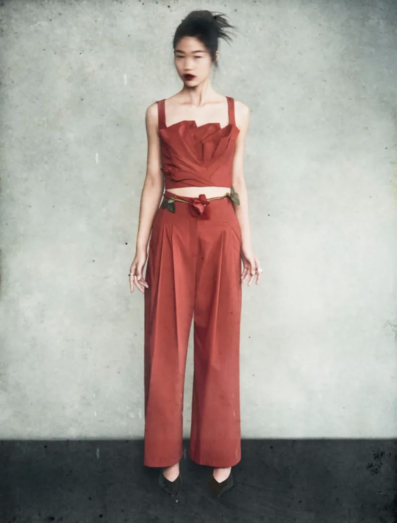 Full-length shot of a woman in an orange two-piece outfit with a floral belt posing in front of a textured grey background. Fashion editorial.